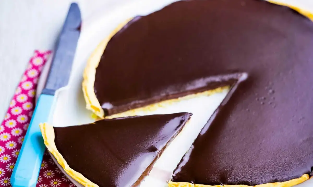 Une part de la meilleure recette de tarte au chocolat, avec une ganache brillante sur une pâte dorée. Un couteau bleu est à côté.