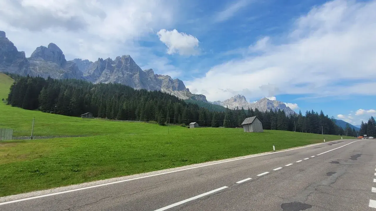 Strada panoramica verso il passo Monte Croce, con prati verdi, boschi di conifere e imponenti vette rocciose sotto un cielo azzurro con nuvole bianche.