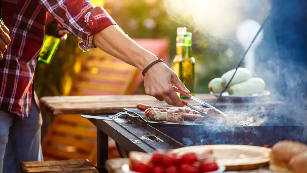 Hombre asa salchichas envueltas en tocino, ideal para saber qué llevar a una barbacoa para sorprender. Hay cervezas y tomates en la mesa.