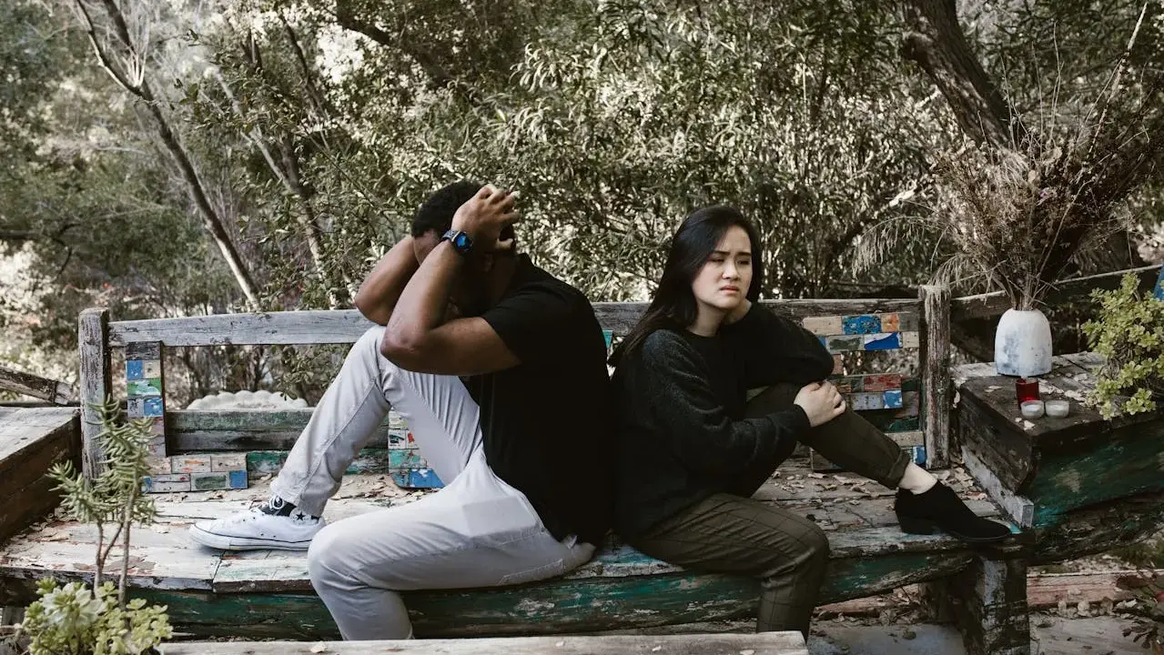 Pareja sentada de espaldas en un banco, con expresiones de angustia. El hombre se agarra la cabeza, ella mira al vacío. Un **vínculo traumático** parece separarlos en este entorno natural.