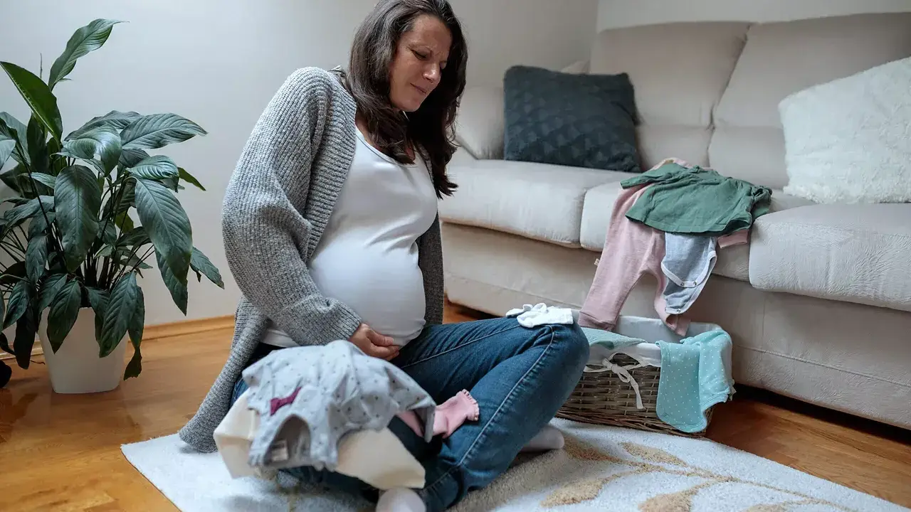 Schwangere Frau sitzt auf dem Boden, hält ihren Bauch und scheint Braxton Hicks Kontraktionen zu spüren, während sie Babykleidung sortiert.