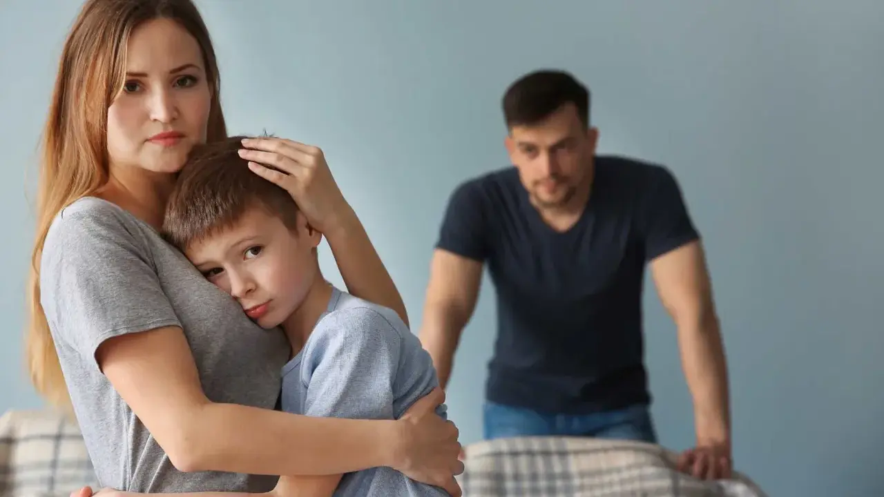 Smutna kobieta tuli syna, podczas gdy partner patrzy z dystansu. Trudno jej zaakceptować dziecko partnera.