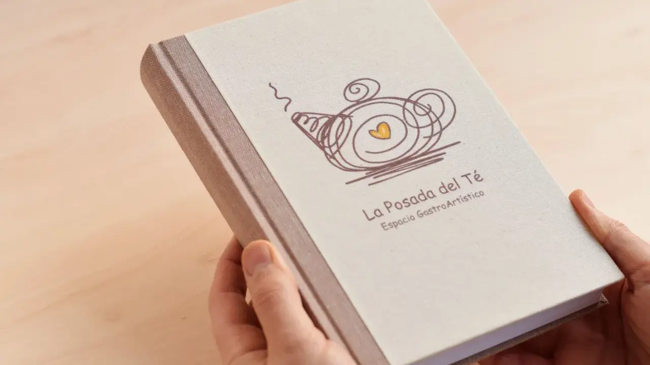 Manos sostienen un libro con tapa de tela, con un diseño de tetera y el texto "La Posada del Té". Una idea para como encuadernar un trabajo de forma original.