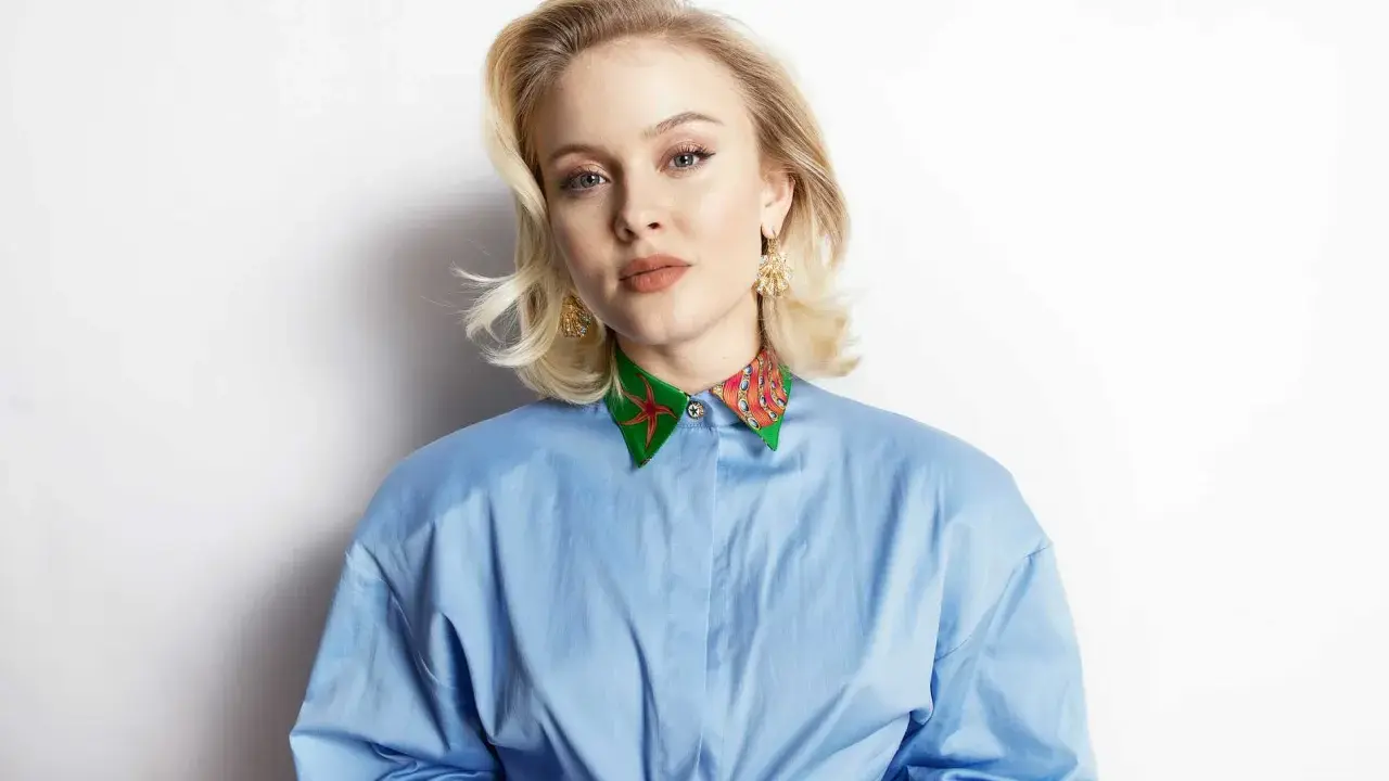 Zara Larsson, känd för sin musik, bär en blå skjorta med en färgglad krage. Hennes skivbolag har säkert hjälpt henne nå framgång.