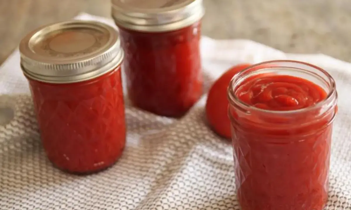 Trois bocaux de ketchup maison, un rouge vif, reposent sur une serviette à motifs. Un tomate entière est visible.