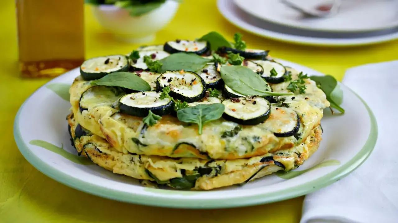 Deliciosa tortilla de calabacín apilada, adornada con rodajas de calabacín, espinacas y perejil fresco.