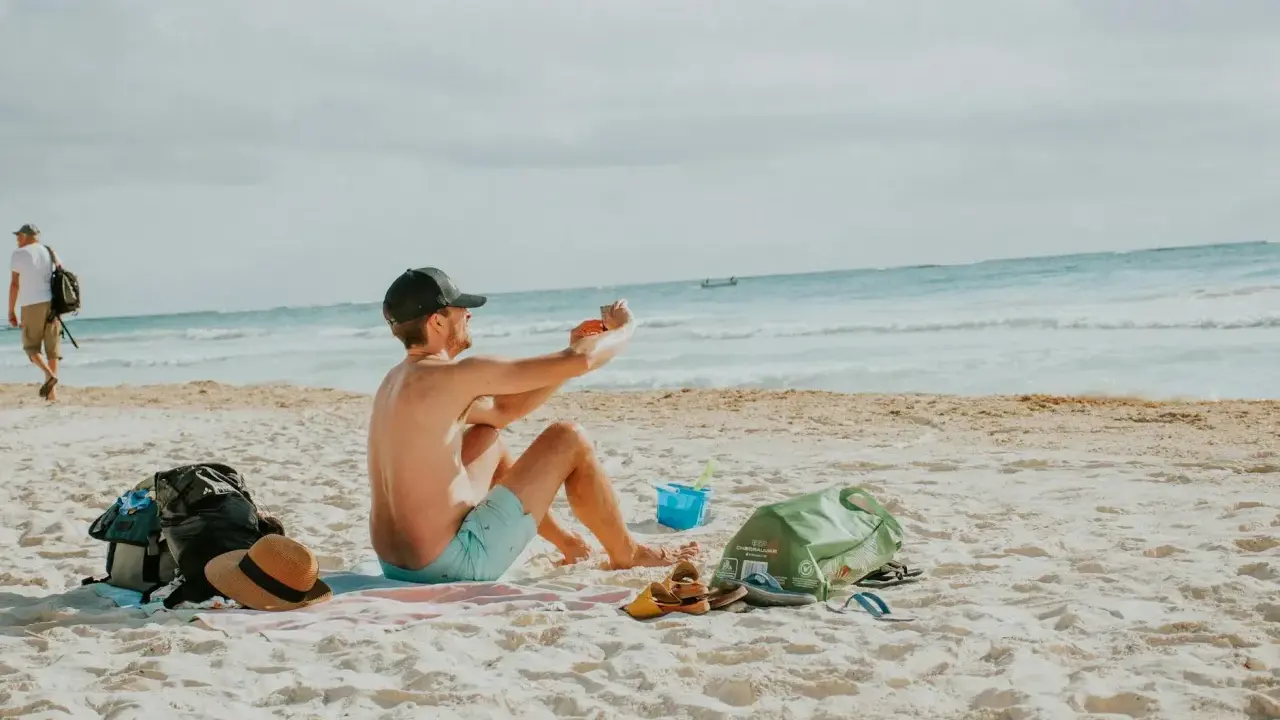 Mężczyzna robi zdjęcie telefonem na plaży, marząc o najkrótszych lotach z Polski na wakacje. Obok leżą torba, kapelusz i sandały.