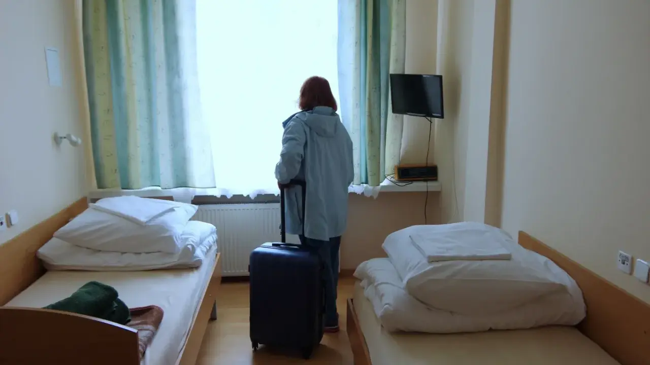 Kobieta z walizką przy oknie, gotowa by dowiedzieć się, jak wyjechać do sanatorium. Pokój z dwoma łóżkami.