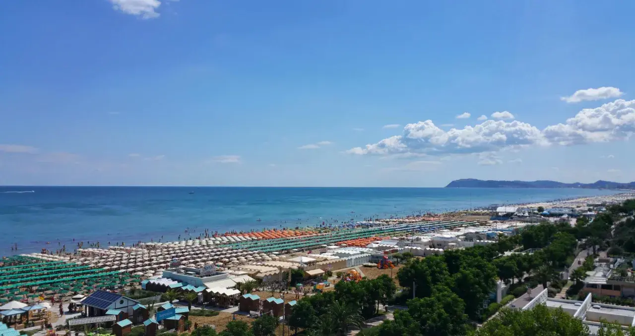 Ampia spiaggia di Riccione con file ordinate di ombrelloni e lettini, bagnanti che si godono il sole e l'acqua cristallina del mare Adriatico sotto un cielo azzurro.