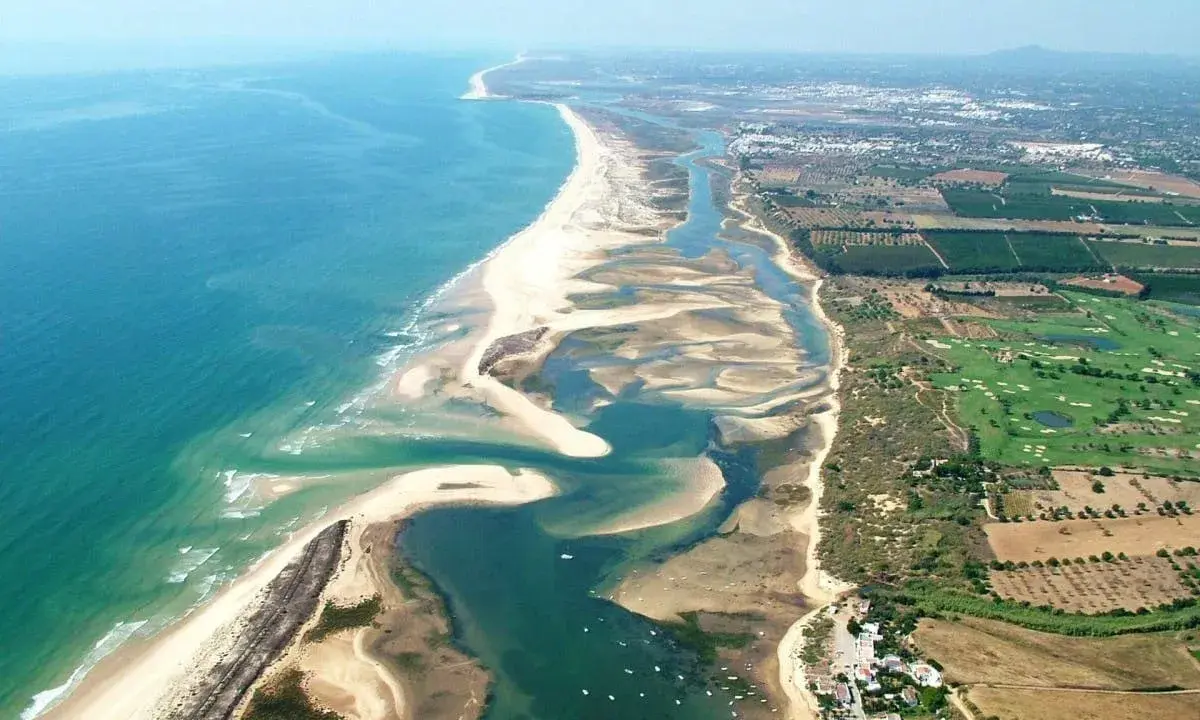 Luftaufnahme der Ria Formosa mit ihren Sandbänken, Lagunen und dem Meer. Im Hintergrund sind Felder und ein Golfplatz zu sehen.