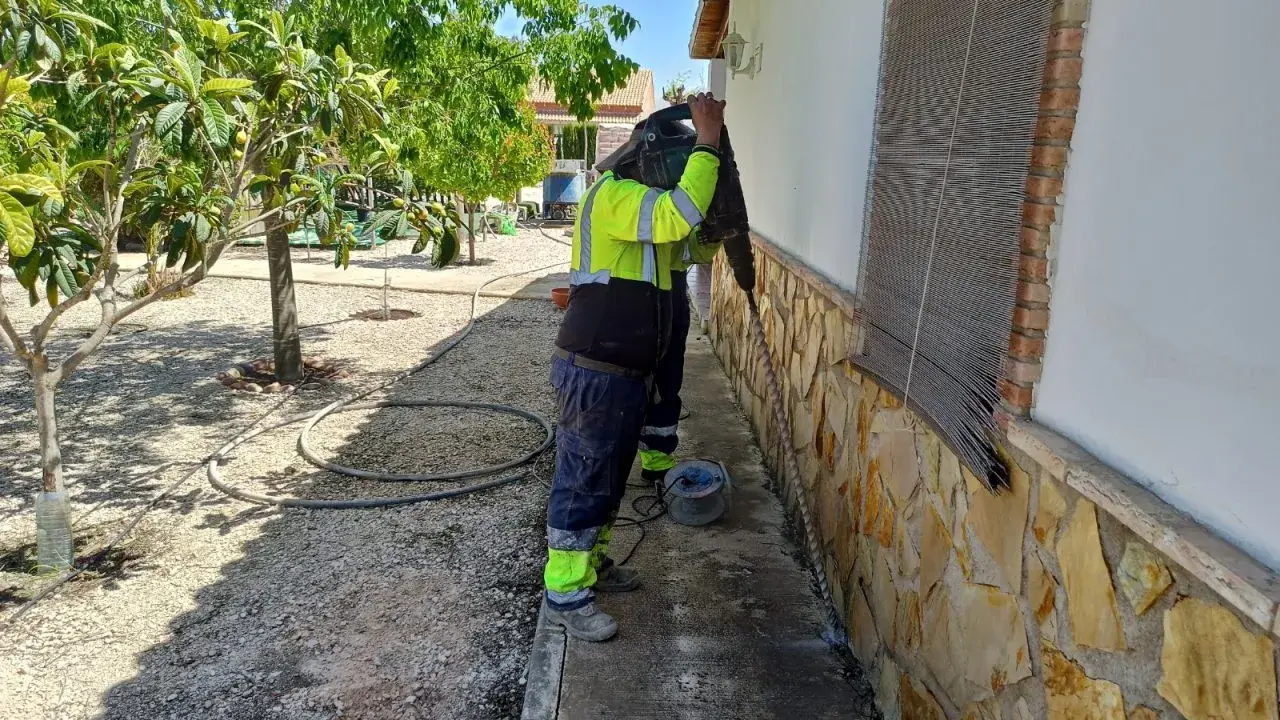 Trabajador con equipo de protección realiza trabajos de inyección de cemento en la base de una pared exterior, rodeado de vegetación.