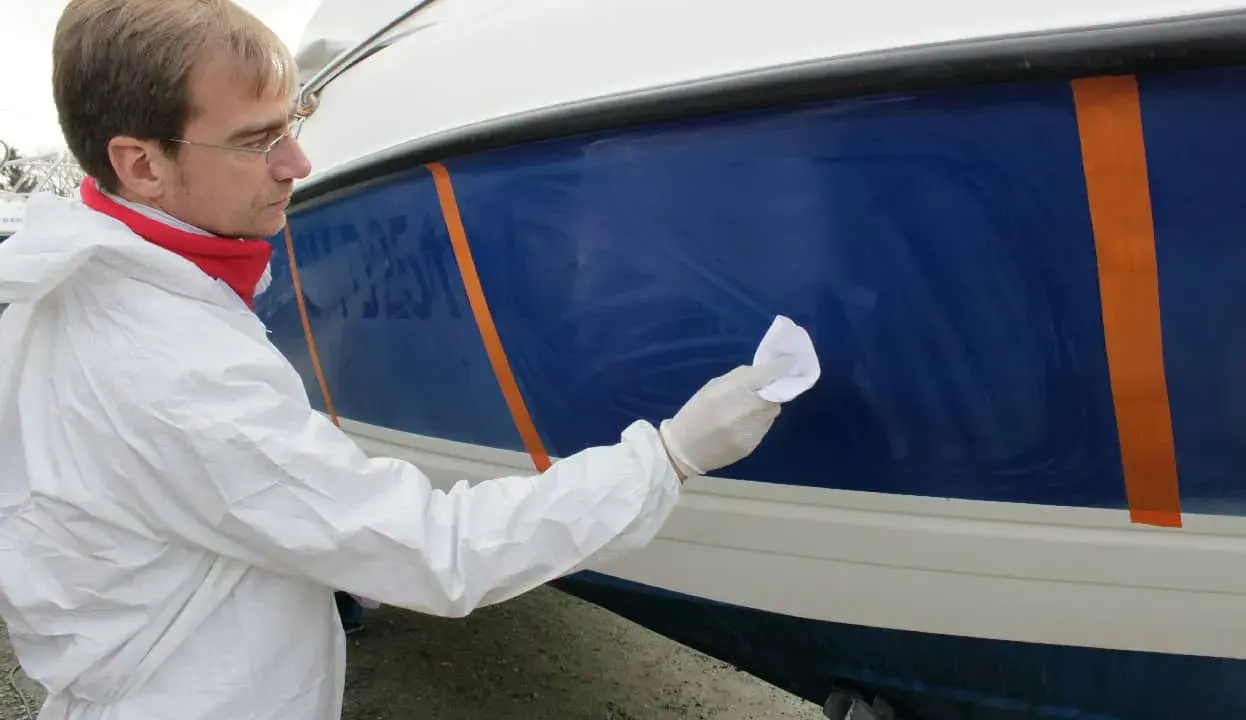 Un homme en combinaison blanche effectue la mise en cire d'un bateau bleu, protégeant la coque pour l'hiver.