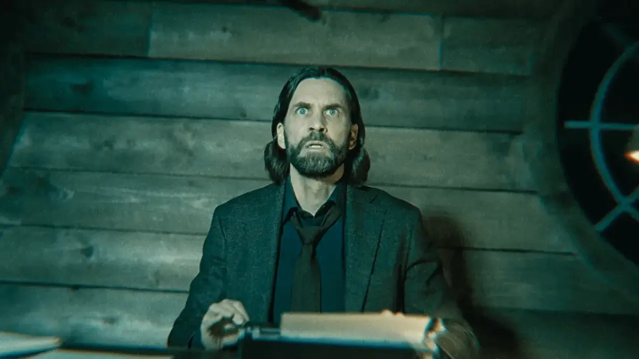 Alan Wake 2: un hombre con barba y pelo largo, con expresión de sorpresa, sentado frente a una máquina de escribir en una habitación de madera.