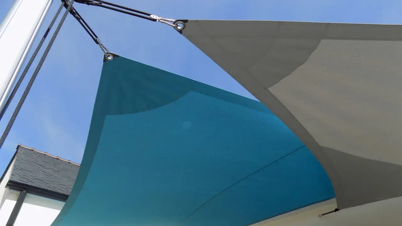 Comment installer un voile d'ombrage sur terrasse : deux voiles, bleu et gris, fixés par des cordages et des poulies à un mât et à la maison.