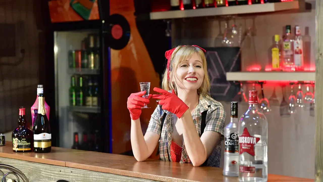Uśmiechnięta barmanka w czerwonych rękawiczkach trzyma kieliszek. Na ladzie stoi duża butelka wódki, idealna na pół litra na dwóch.