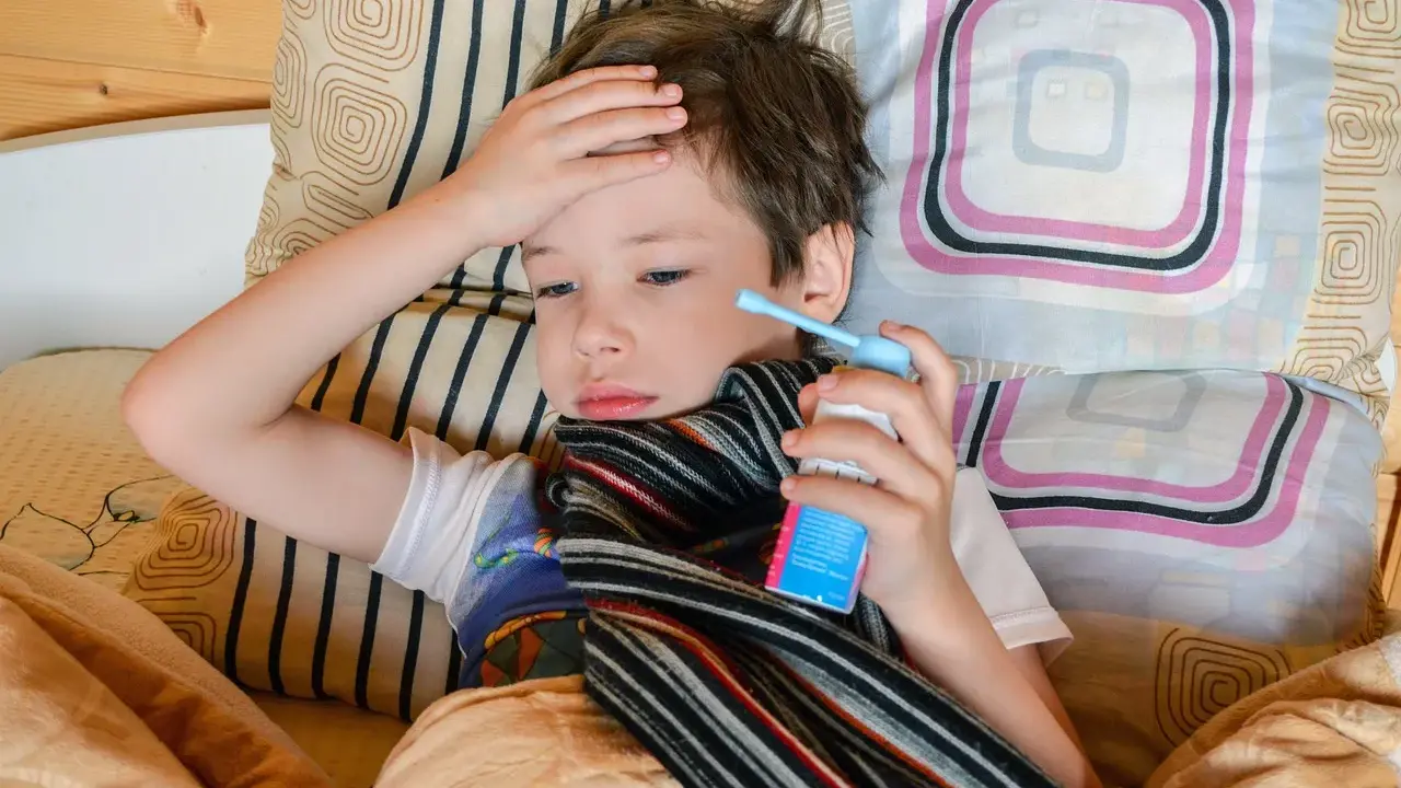 Chory chłopiec leży w łóżku, trzymając w dłoni inhalator. Jego ręka spoczywa na czole, a na szyi ma szalik. To mogą być objawy astmy.