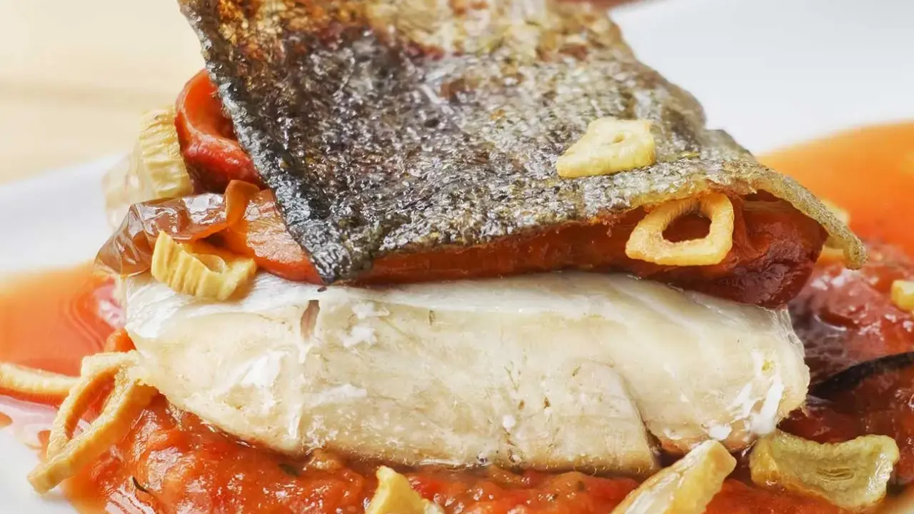 Bacalao a la riojana con piel crujiente, pimientos y ajo frito, servido sobre una cama de salsa de tomate.