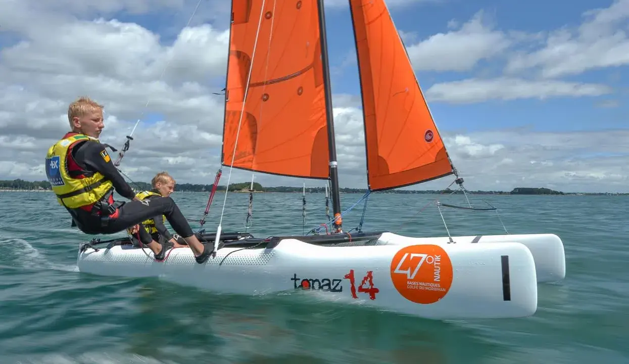 Deux jeunes garçons naviguent sur un petit voilier 2 places, leurs voiles oranges gonflées par le vent sous un ciel bleu et nuageux.