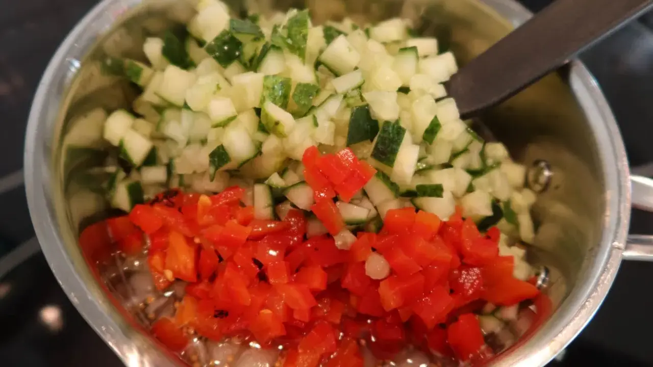 Finhackad gurka och röd paprika i en kastrull, redo för ett bostongurka recept. En sked vilar i blandningen.