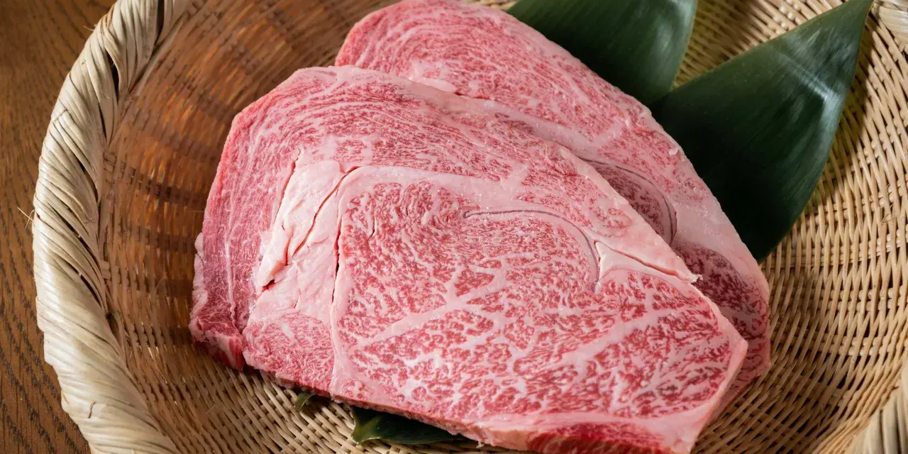 Dos cortes de carne japonesa cara, con marmoleado intenso, reposan sobre hojas verdes en una cesta de mimbre.