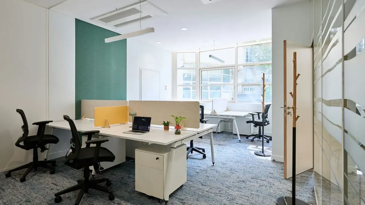 Un bureau chaleureux avec des postes de travail modernes, des cloisons acoustiques et un tapis bleu doux. L'espace est lumineux grâce aux grandes fenêtres.