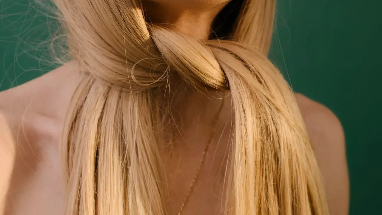 Blondes, gesundes Haar, das sich um den Hals schlingt. Ein zarter goldener Anhänger ist unter den Haaren zu sehen.