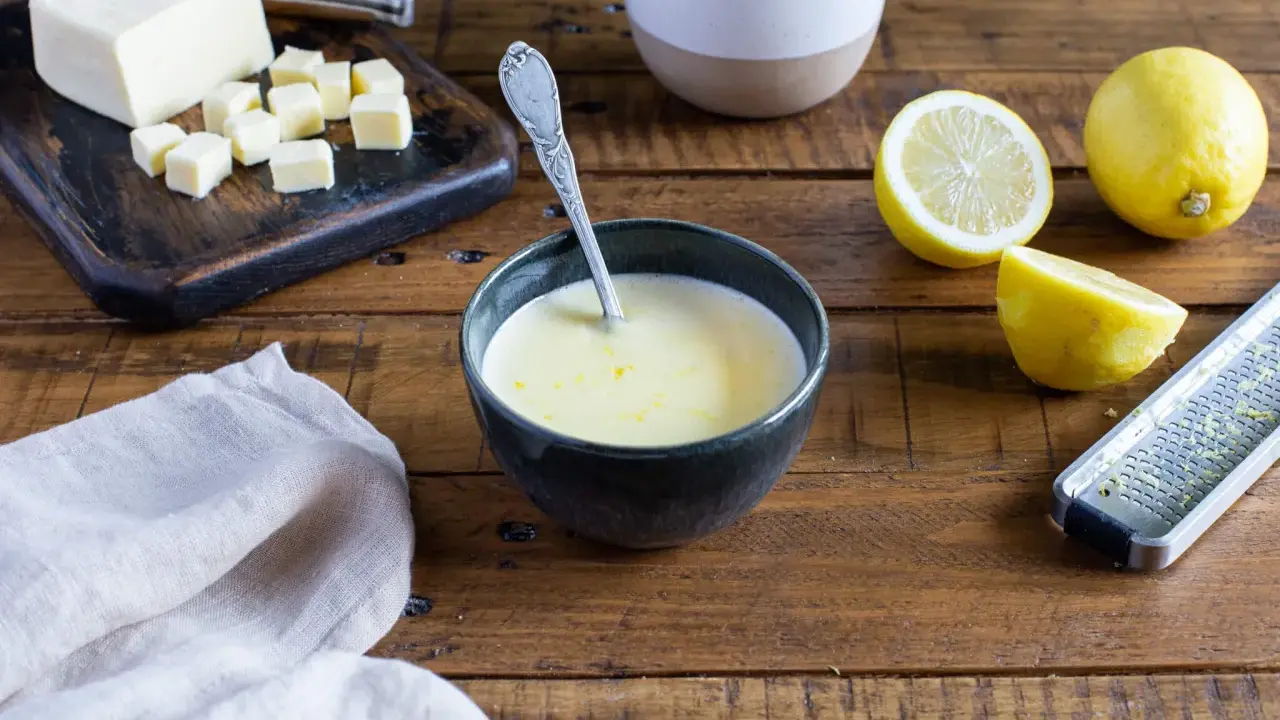 Un bol de sauce beurre citron, avec des citrons, du beurre coupé en dés et une râpe à zester sur une table en bois.