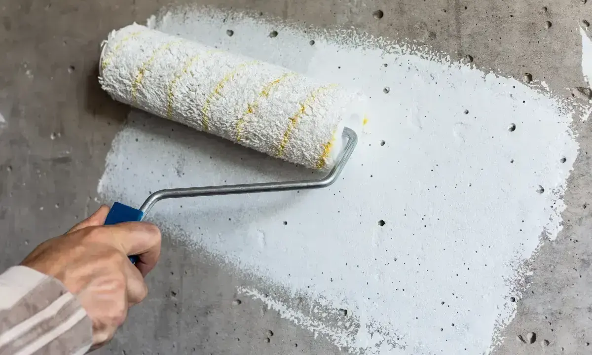 Ręka z wałkiem nakłada biały grunt na betonową ścianę. To pierwszy krok, jak gruntować ściany przed malowaniem.