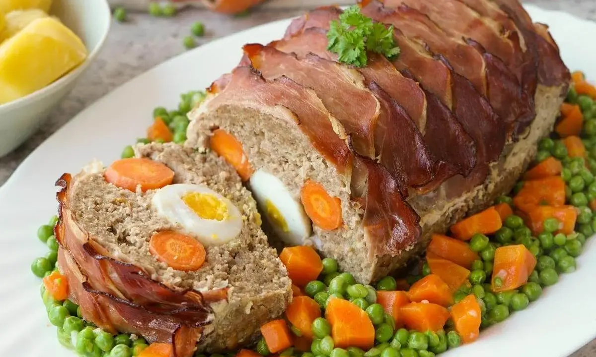 Ein saftiger falscher Hase, umwickelt mit Speck, gefüllt mit Eiern und Karotten, serviert auf Erbsen und Karotten.