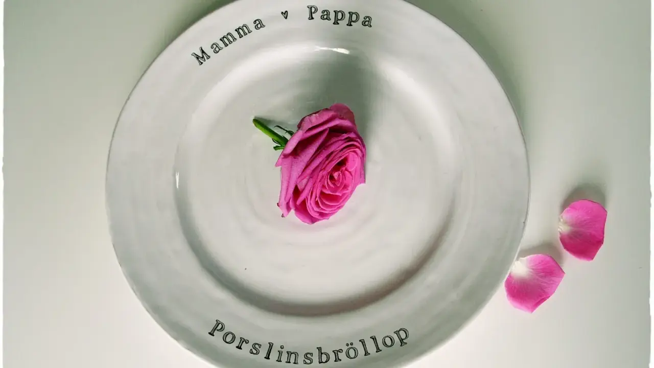 En vit tallrik med texten "Mamma ♡ Pappa" och "Porslinsbröllop". En rosa ros ligger i mitten. Ett vackert minne för de som varit gifta i 20 år.