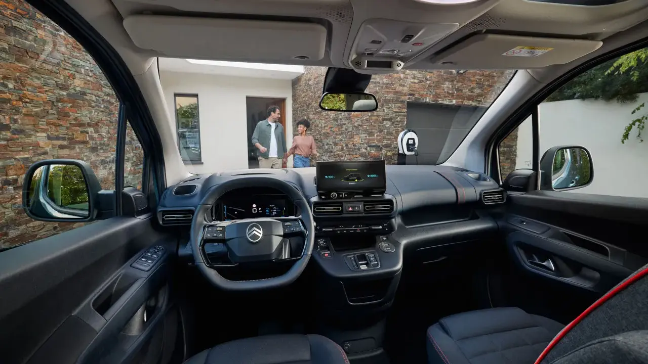 Interior de la nueva Citroën Berlingo, con pantalla táctil, volante moderno y asientos cómodos. Afuera, una pareja se acerca a un punto de carga.