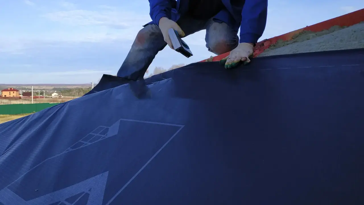 Un ouvrier en bleu, ganté, utilise un outil pour la pose d'un pare pluie sur un toit. Le ciel est bleu avec quelques nuages.