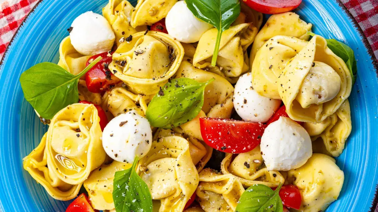 Ein bunter Tortellini Salat mit Mozzarella-Kugeln, Tomatenstücken und frischem Basilikum, serviert auf einem blauen Teller.