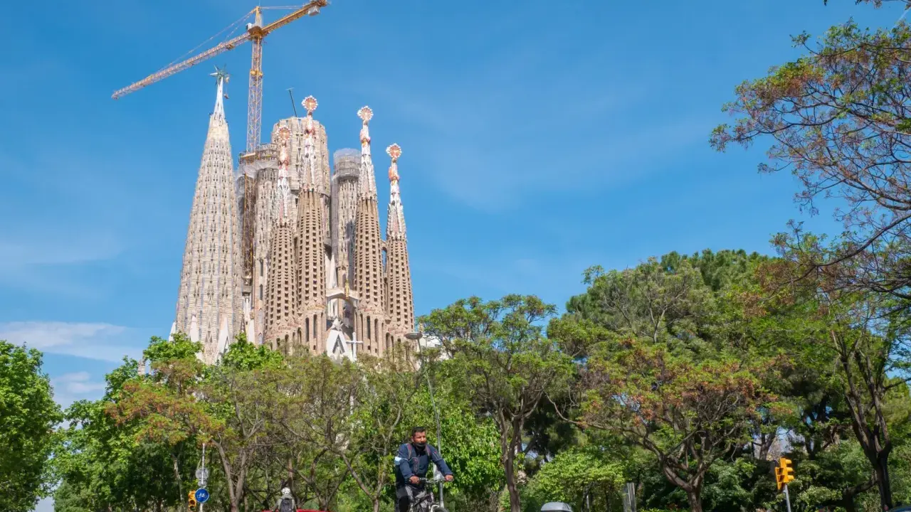 Sagrada Familia w Barcelonie, wciąż w budowie, góruje nad zielonym parkiem. Na ulicy widać rowerzystę i samochody.
