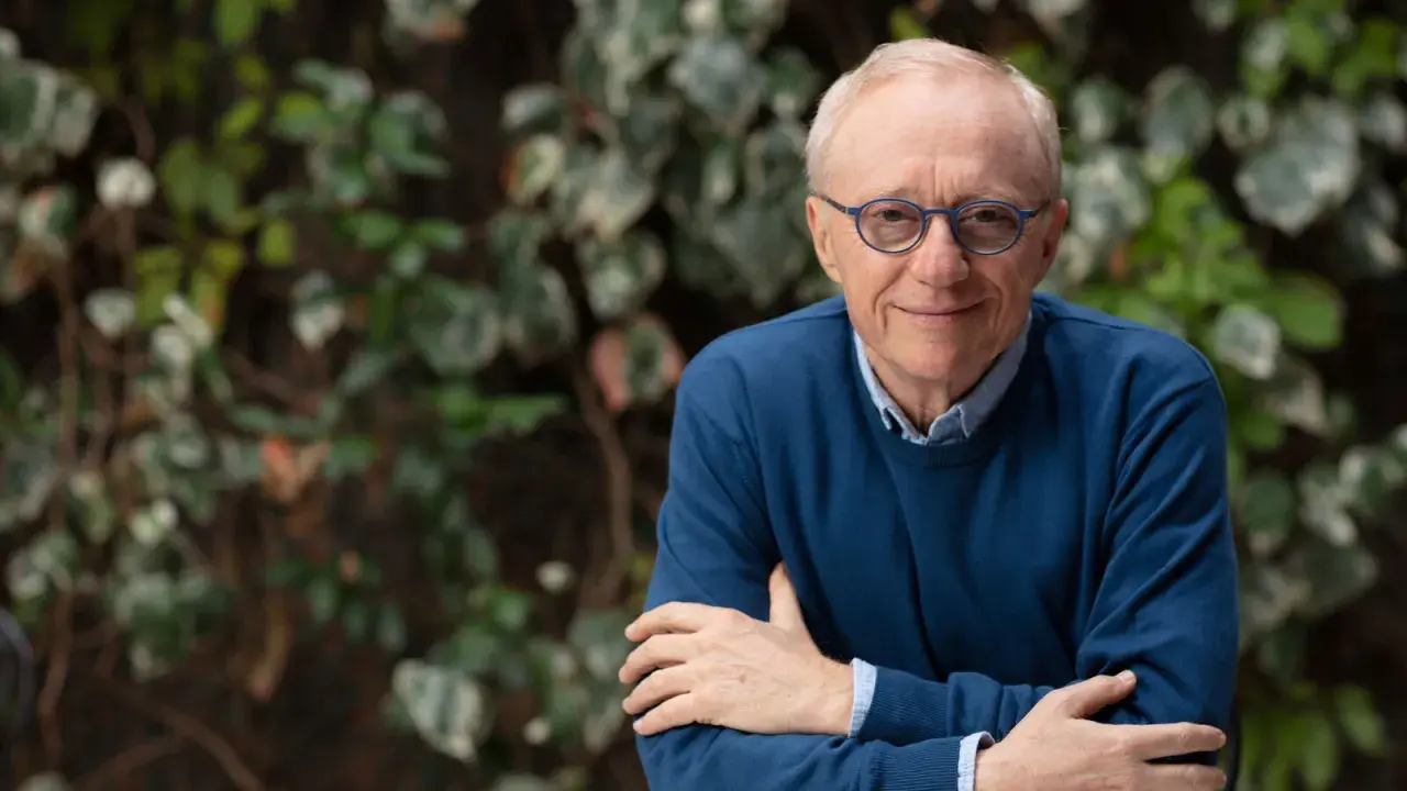 David Grossman, con occhiali blu e maglione scuro, sorride con le braccia incrociate su sfondo verde.