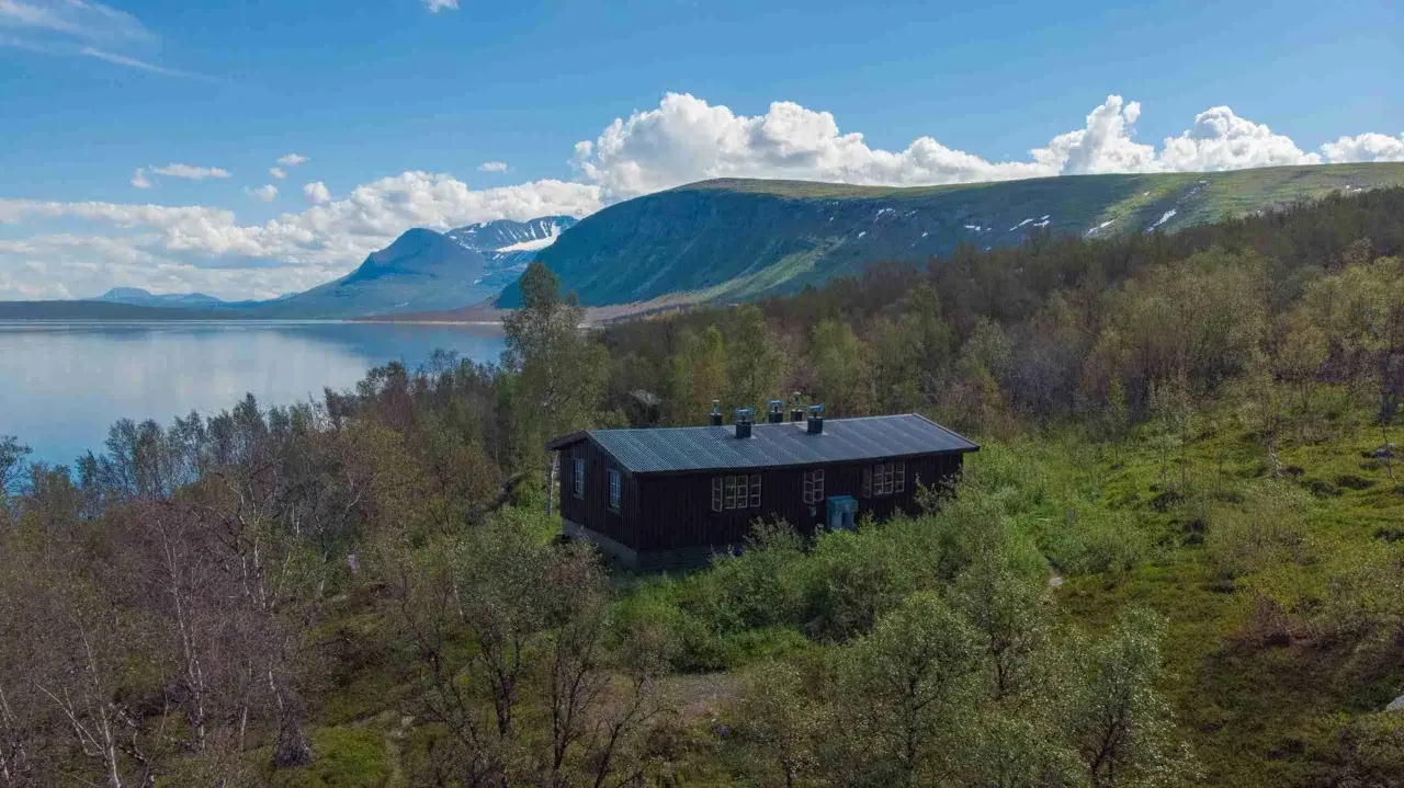 Mörkbrun byggnad vid en sjö, omgiven av björkskog och med fjäll i bakgrunden. En idyllisk plats för ritsem camping.