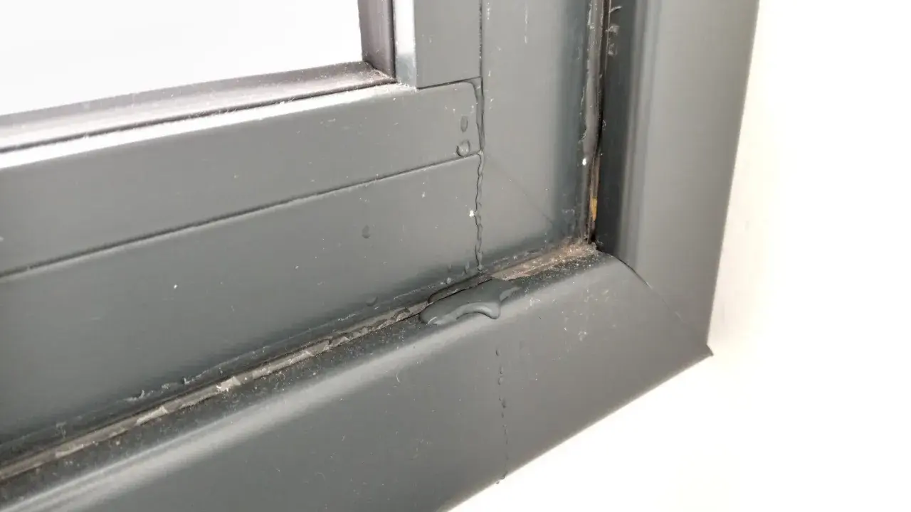Gotas de lluvia resbalan por el marco de una ventana gris, dejando un rastro húmedo.