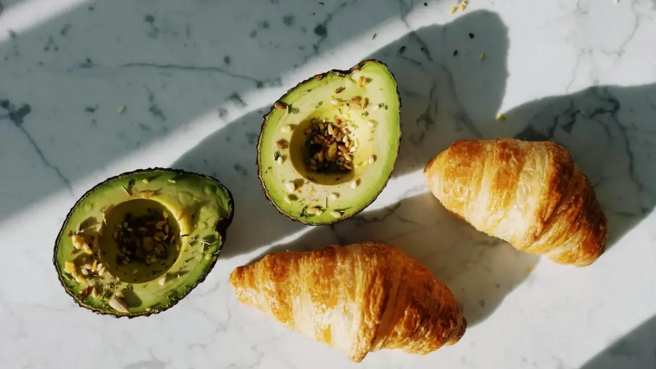 Dwa połówki awokado z nasionami i dwa chrupiące croissanty na marmurowym blacie. Idealne składniki na pyszne awokado przepisy.