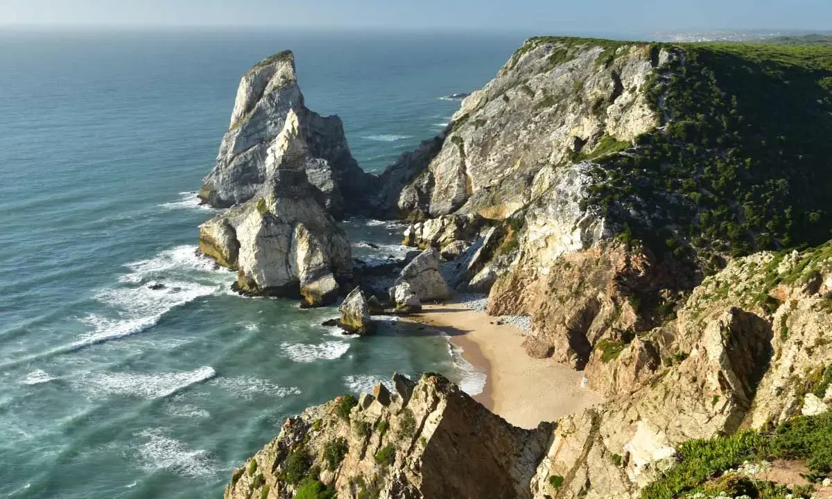 Steile Felsklippen umgeben einen einsamen sintra strand. Das türkisfarbene Meer bricht sich an den Felsen.