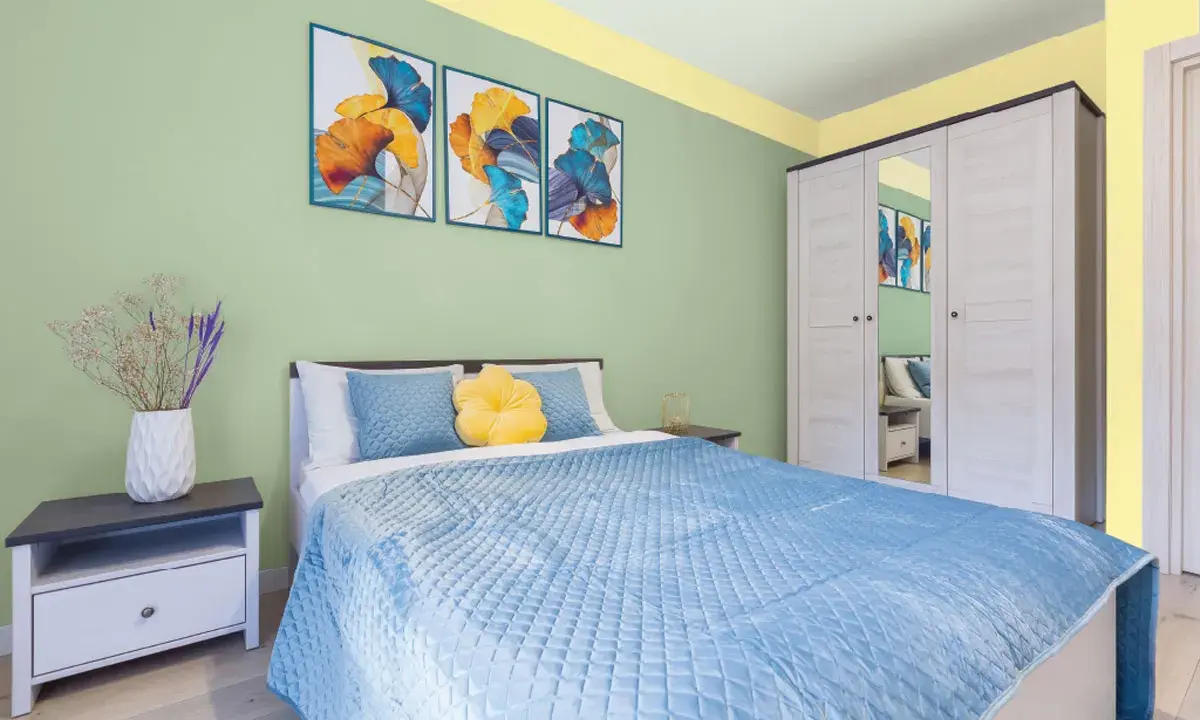 Dormitorio con paredes verde menta y detalles amarillos en el techo. Cama azul, mesita de noche y armario blanco. Inspiración de colores para pintar un dormitorio pequeño.