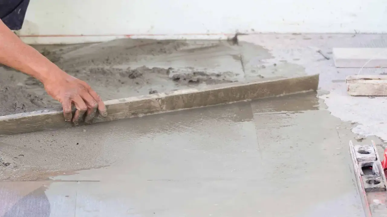 Ręka wyrównuje świeżą betonową wylewkę na ziemi za pomocą łaty. Kluczowe etapy, jak zrobić wylewkę betonową na ziemi.