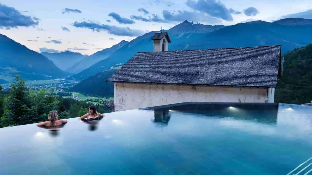 Due donne in una piscina a sfioro ammirano il panorama di Bormio e dintorni, con montagne maestose e un piccolo edificio storico.