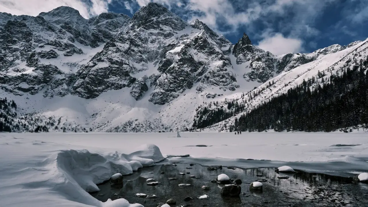 Morskie Oko zimową porą. Lodowate wody jeziora odbijają ośnieżone szczyty Tatr, a na horyzoncie widać ludzi spacerujących po zamarzniętej tafli.