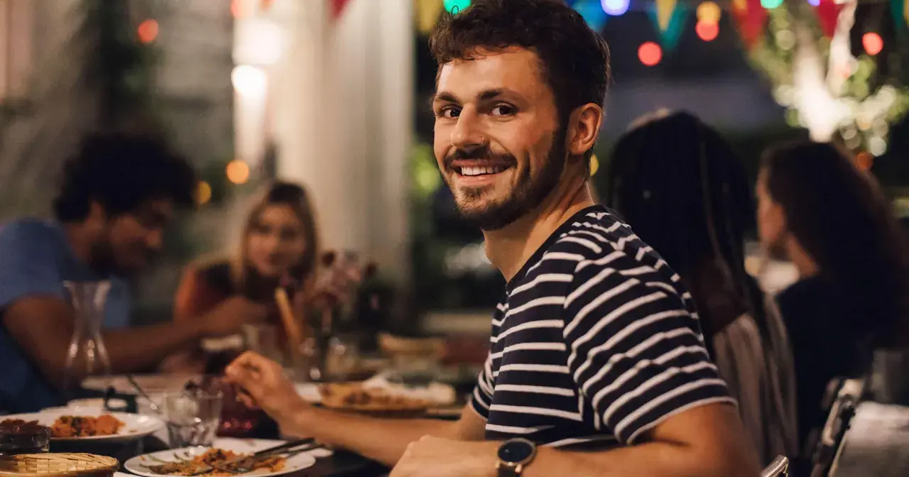 Uśmiechnięty mężczyzna na tle przyjaciół przy stole. Idealny, tani pomysł na wieczór kawalerski z jedzeniem i dobrym towarzystwem.