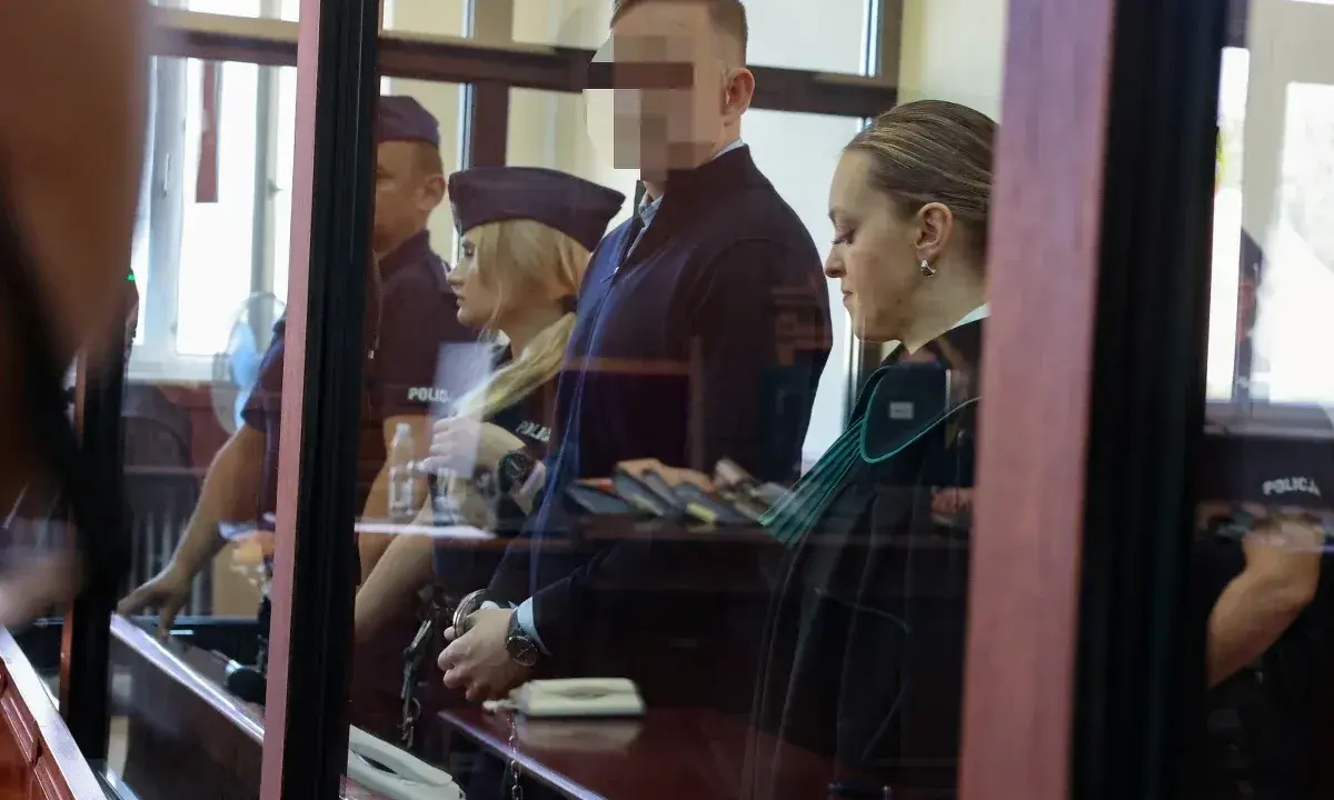 Osoba w kajdankach, prawniczka i policjanci w sali sądowej.