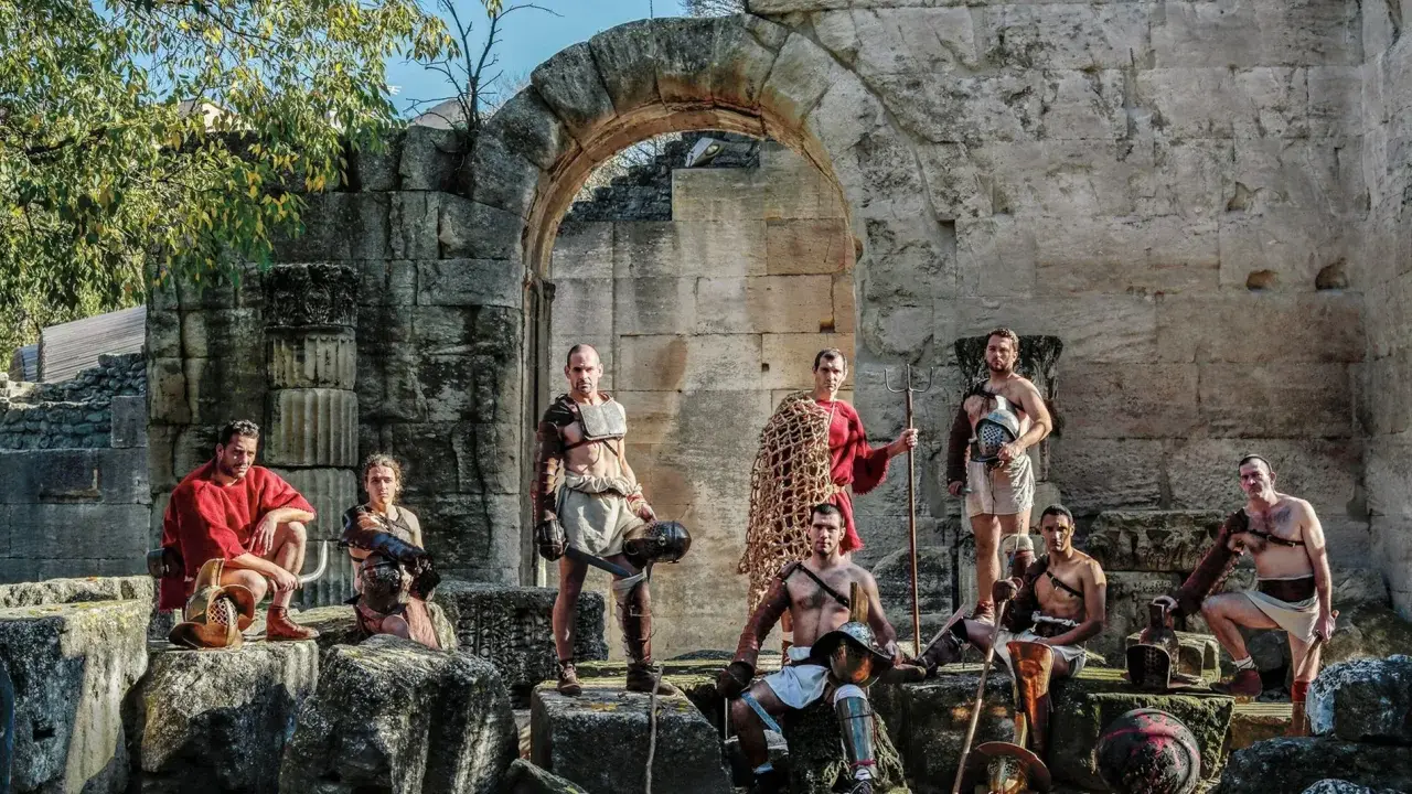 Un groupe de guerriers, prêts à combattre, pose devant des ruines antiques. Ces hommes, tels des gladiateurs romains, arborent armures et armes, prêts pour l'arène.