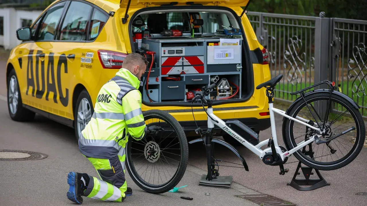 ADAC-Mitarbeiter repariert eine Fahrradpanne. Ein gelbes ADAC-Auto mit Werkzeugkasten steht daneben.