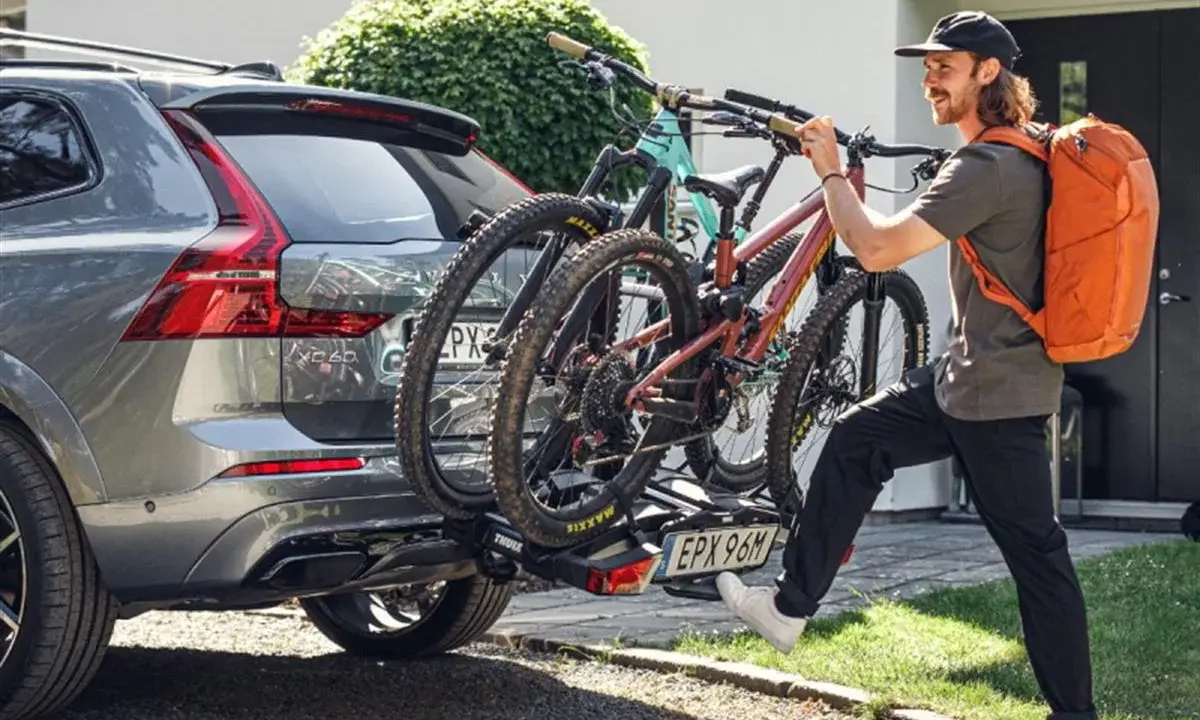 Mężczyzna z pomarańczowym plecakiem montuje bagażnik rowerowy na hak do Volvo XC60. Dwa rowery górskie są już zamocowane. Przepisy dotyczące bagażnika rowerowego na hak są kluczowe dla bezpieczeństwa.