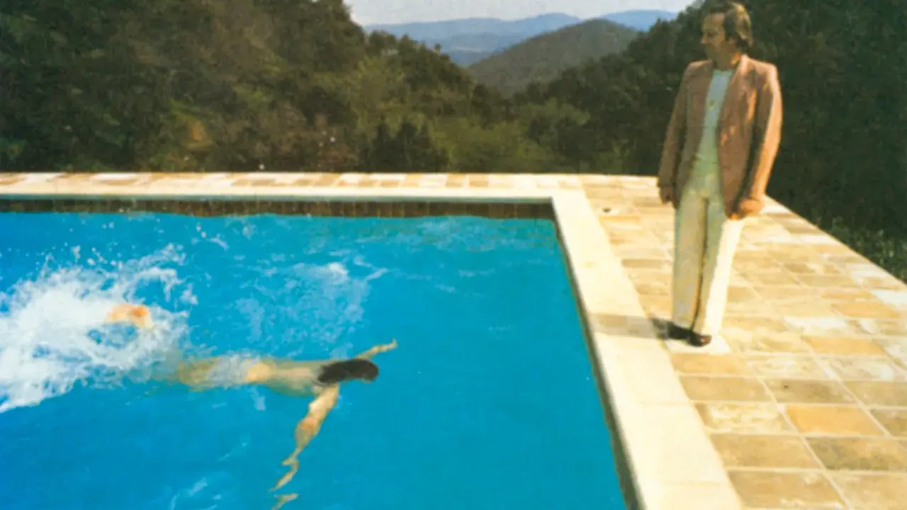 A portrait of an artist by a pool with two figures. One figure swims in the bright blue water, while another stands on the tiled edge, looking out at the mountainous landscape.