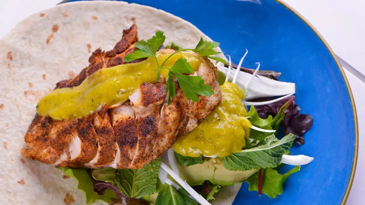 Delicioso wrap de pollo con aguacate, lechugas variadas y salsa verde, servido en un plato azul vibrante.